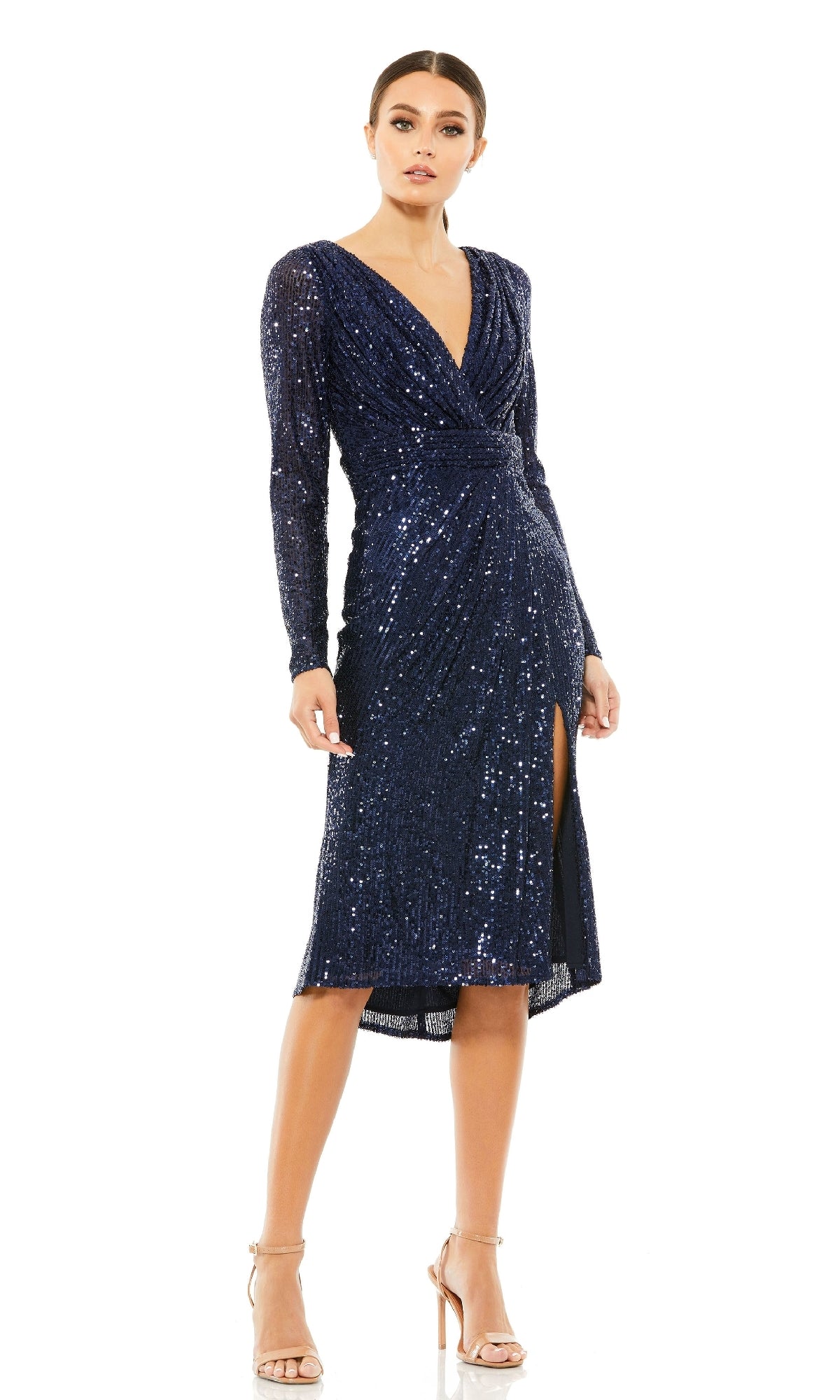 A woman wears the Mac Duggal 26555 long sleeve sequin party dress in navy blue with a V-neckline, knee-length hem, and front slit, styled with nude high-heeled sandals.